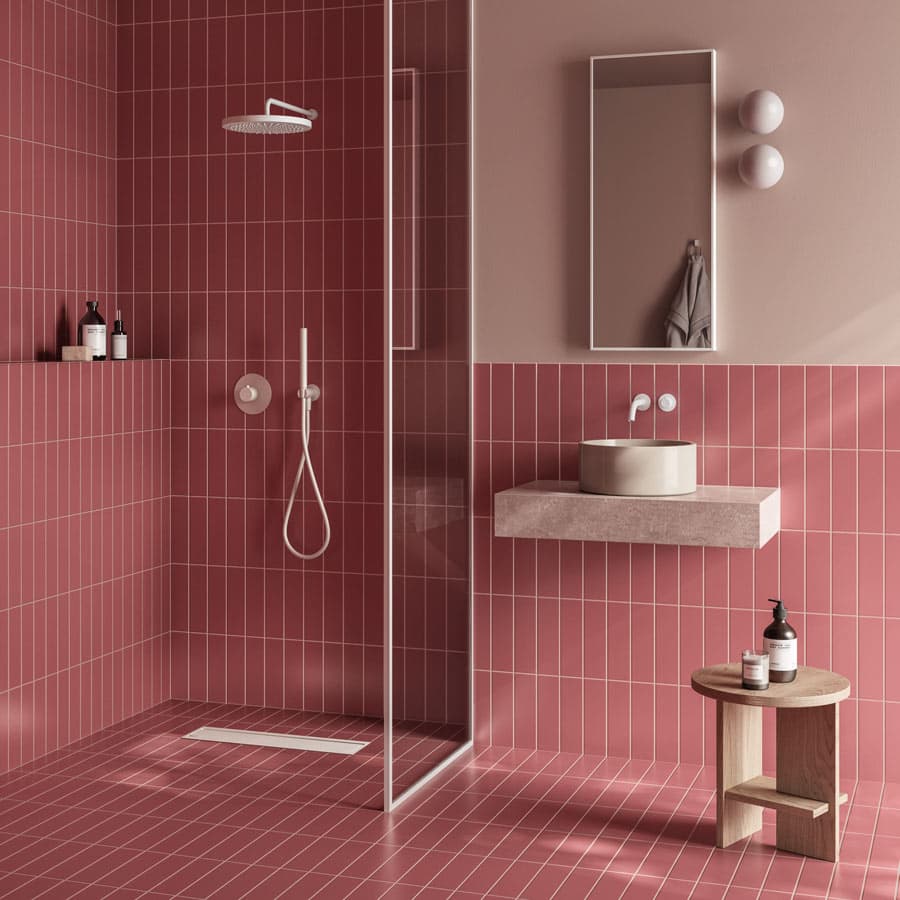Modern bathroom with pink tiles, a glass shower, round mirror, wall-mounted sink, and wooden stool with toiletries.