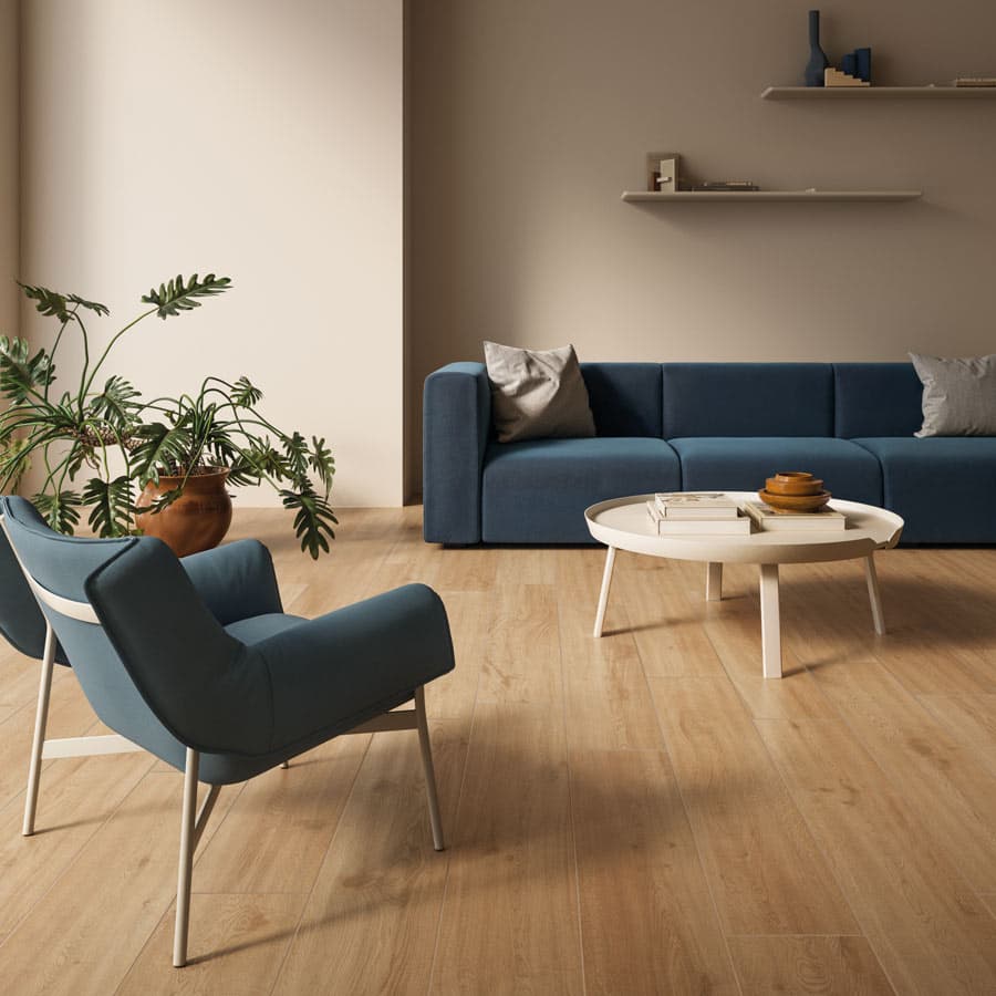 Minimalist living room with a blue sofa, matching armchair, round white coffee table, wooden floor, and a potted plant. Neutral wall with shelves.