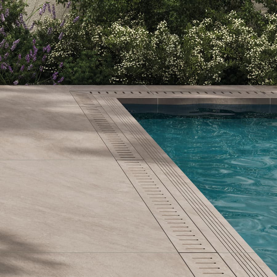 A corner of a swimming pool with clear blue water, surrounded by beige tiles and lush greenery in the background.